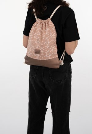 Person trägt ein schwarzes Hemd und schwarze Hose mit einem beige-braun gemusterten Rucksack mit Kordelzug auf dem Rücken vor weißem Hintergrund.