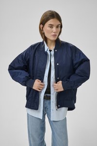 Junge Frau, die eine marineblaue Bomberjacke über einem hellblauen Hemd und hellblauen Jeans gegen einen schlichten grauen Hintergrund anpasst.
