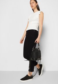 Top branco canelado com bordas de mangas frufru, combinado com calças pretas com detalhes de botões. Modelo segurando uma bolsa preta em formato de balde e usando ténis pretos.