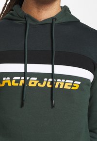 Mikina s kapucí v tmavě zelené barvě s vodorovnými pruhy černé, bílé a žluté. Obsahuje logo "JACK&JONES" ve výrazných kontrastních barvách.