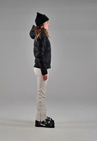 Woman wearing a black beanie, black puffer jacket, beige pants, and black ski boots standing in profile against a gray background.