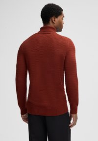 Uomo visto di spalle che indossa un maglione a collo alto in knit testurizzato color ruggine e pantaloni neri, su uno sfondo grigio uniforme.