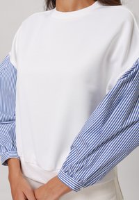 Vit sweatshirt med blå och vita randiga puffärmar, rund halsringning och ribbad nederkant; kombinerar mjukt tyg och en strukturerad design.