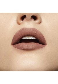 Rouge à lèvres nude mat sur des lèvres pulpeuses, montrant une texture lisse et des contours définis. Un teint clair en arrière-plan révèle un éclat naturel.