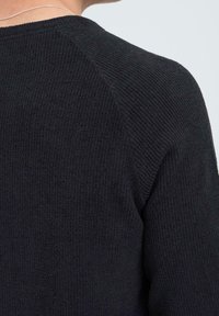 Texturiertes schwarzes Sweatshirt mit einem gerippten Muster, Raglanärmeln und einem subtilen Glanz, das einen Nahaufnahme des Schulterbereichs zeigt.