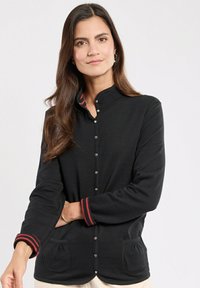 Schwarzes Langarmhemd mit Stehkragen, rot gestreiften Manschetten, Druckknöpfen und Taschen vorne. Weicher Stoff mit glatter Textur.