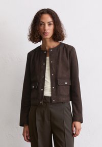Braune Wildlederjacke mit rundem Ausschnitt, Druckknopfverschluss und zwei Vordertaschen. Getragen über einem hellen Oberteil und dunklen Hosen.