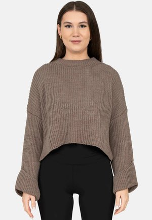 Vrouw met lang bruin haar, die gouden creolen draagt, een bruin geribd cropped truitje met wijde mouwen en een zwarte broek.