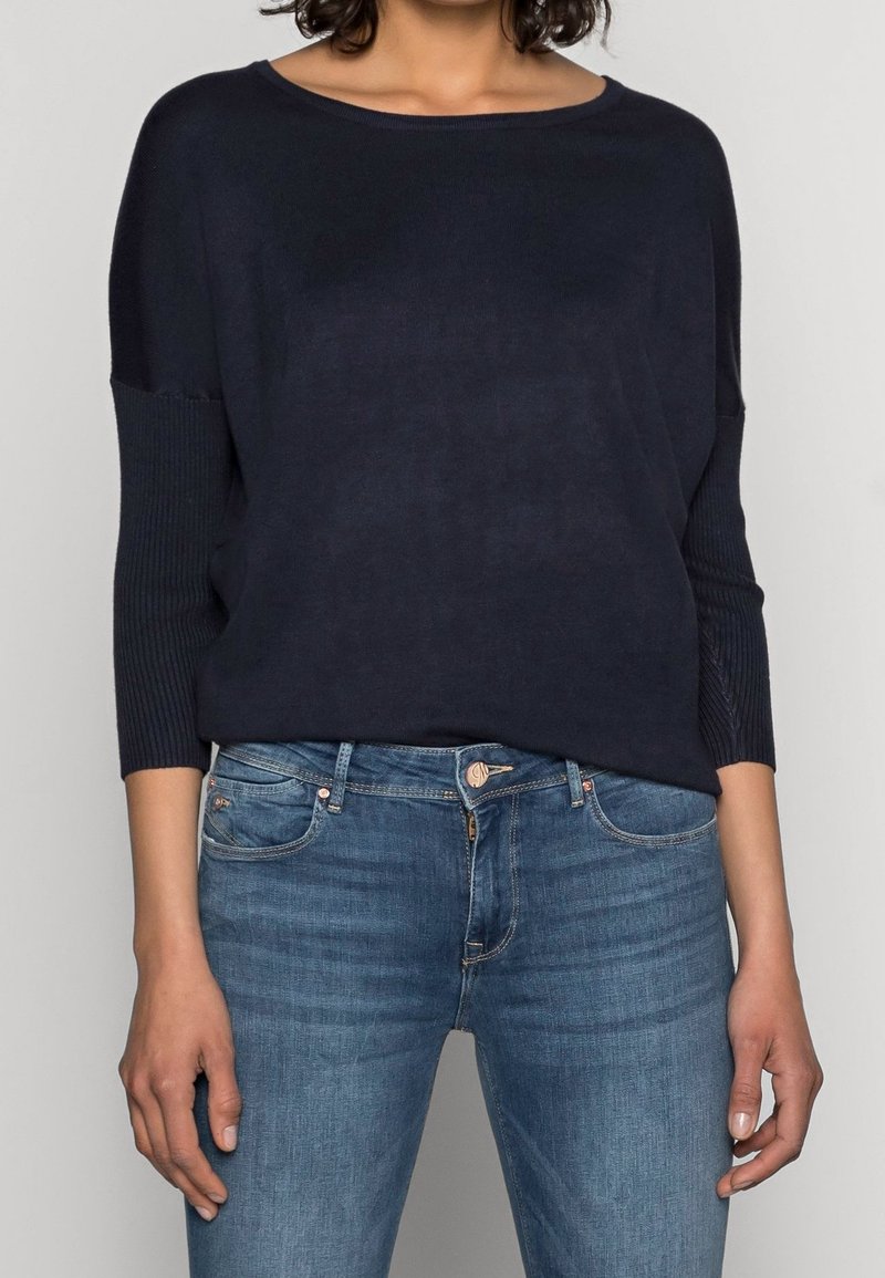 Mörkblå stickad tröja med avslappnad passform, ribbade ärmar och mjuk textur, i kombination med ljusblå tajta jeans.