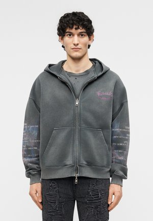 Jeune homme aux cheveux bouclés portant un sweat à capuche gris zippé avec du texte imprimé sur les manches et un jean noir déchiré, debout face à l'avant.