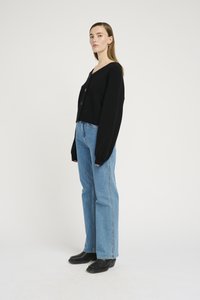 Cardigan preto de malha com decote em V, ajuste oversized e botões frontais, combinado com jeans de ganga azul de cintura alta e sapatos pretos.