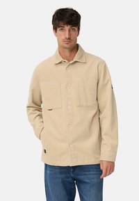 Beige corduroy jas met een knoopsluiting aan de voorkant, twee borstzakken en een losse pasvorm. Heeft een subtiele textuur en een logodetail op de mouw.