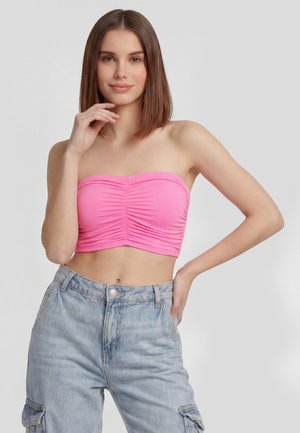 Frau mit glatten braunen Haaren, die ein gerafftes pinkfarbenes trägerloses Crop-Top und hoch taillierte hellblaue Denim-Cargohosen trägt und mit der Hand nahe am Gesicht posiert.