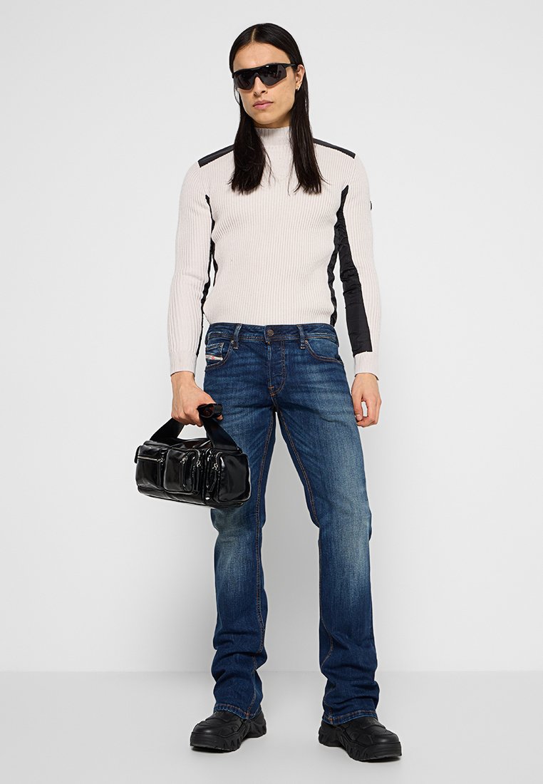 Person, die eine Sonnenbrille, einen weißen gerippten Pullover mit schwarzen Akzenten, blaue Jeans und schwarze Schuhe trägt und eine schwarze Handtasche hält.