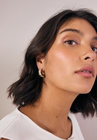 Gold hoop earrings with a textured, ridged design. Worn by a person with dark hair, showcasing a close-up of the earring against a neutral background.
