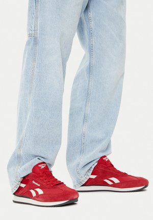 Czerwone sneakersy z zamszu z białymi detalami, zawiązywane na sznurówki i z wyściełanym kołnierzem. Noszone z jasnoniebieskimi dżinsami.