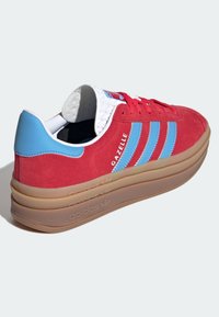 Röd mockasneaker med gummisula, blå detaljer och tre vita ränder. Har en vadderad krage och logotyp på tungan.