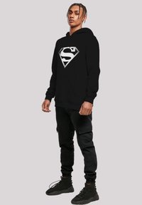 Black hoodie featuring a large white Superman logo on the front. Worn with black cargo pants and black athletic shoes. Textured fabric.