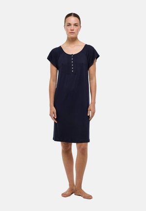 Femme debout pieds nus portant une robe bleu marine au genou avec manches courtes et patte de boutonnage sur un fond clair uni.