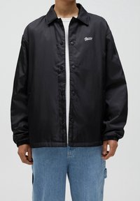 Czarna nylonowa kurtka z guzikami z przodu, kołnierzykiem i elastycznymi mankietami. Posiada małe logo na lewej piersi.