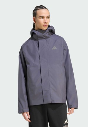 Person med flettet hår iført en løs mørkegrå hættetrøje og sorte Adidas-bukser, der kigger til siden.
