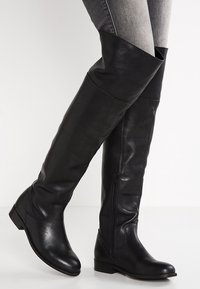 Des bottes en cuir noires montant jusqu'aux genoux avec une fermeture éclair sur le côté, portées sur un jean skinny gris, se tenant devant un fond blanc uni.