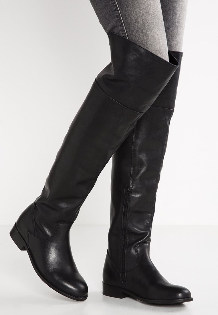 Des bottes en cuir noires montant jusqu'aux genoux avec une fermeture éclair sur le côté, portées sur un jean skinny gris, se tenant devant un fond blanc uni.