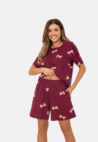 Kobieta uśmiechająca się, ubrana w luźne bordowe szorty oraz pasującą krótki rękaw koszulę w żółte wzory w kokardki na jednolitym jasnym tle.