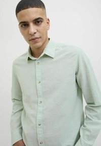 Cămașă verde deschis cu mâneci lungi, realizată dintr-un material texturat, având guler cu nasturi și nasturi vizibili de-a lungul plăcii frontale.
