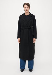 Donna in piedi di fronte, che indossa un lungo cappotto nero con cintura, jeans blu, scarpe nere a punta e orecchini a cerchio argentati.