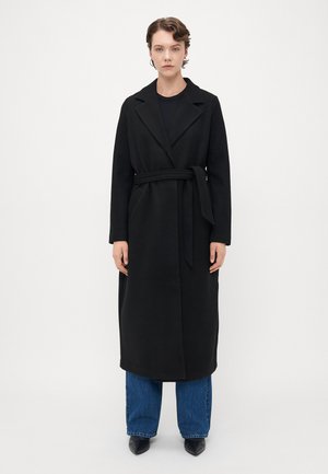Femme se tenant face à l'avant, portant un long manteau noir ceinturé, un jean bleu, des chaussures noires à talons pointus et des boucles d'oreilles en argent.