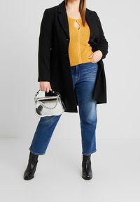 Czarny płaszcz na musztardowym swetrze w prążki z marszczeniem, w połączeniu z niebieskimi dżinsami typu straight-leg i czarnymi botkami za kostkę. Akcent w postaci srebrnej torebki.