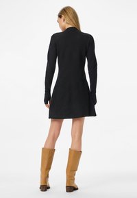 Žena otočená chrbtom má na sebe čierne šaty s dlhým rukávom a hnedé kožené čižmy po kolená na jednoduchom bielom pozadí.