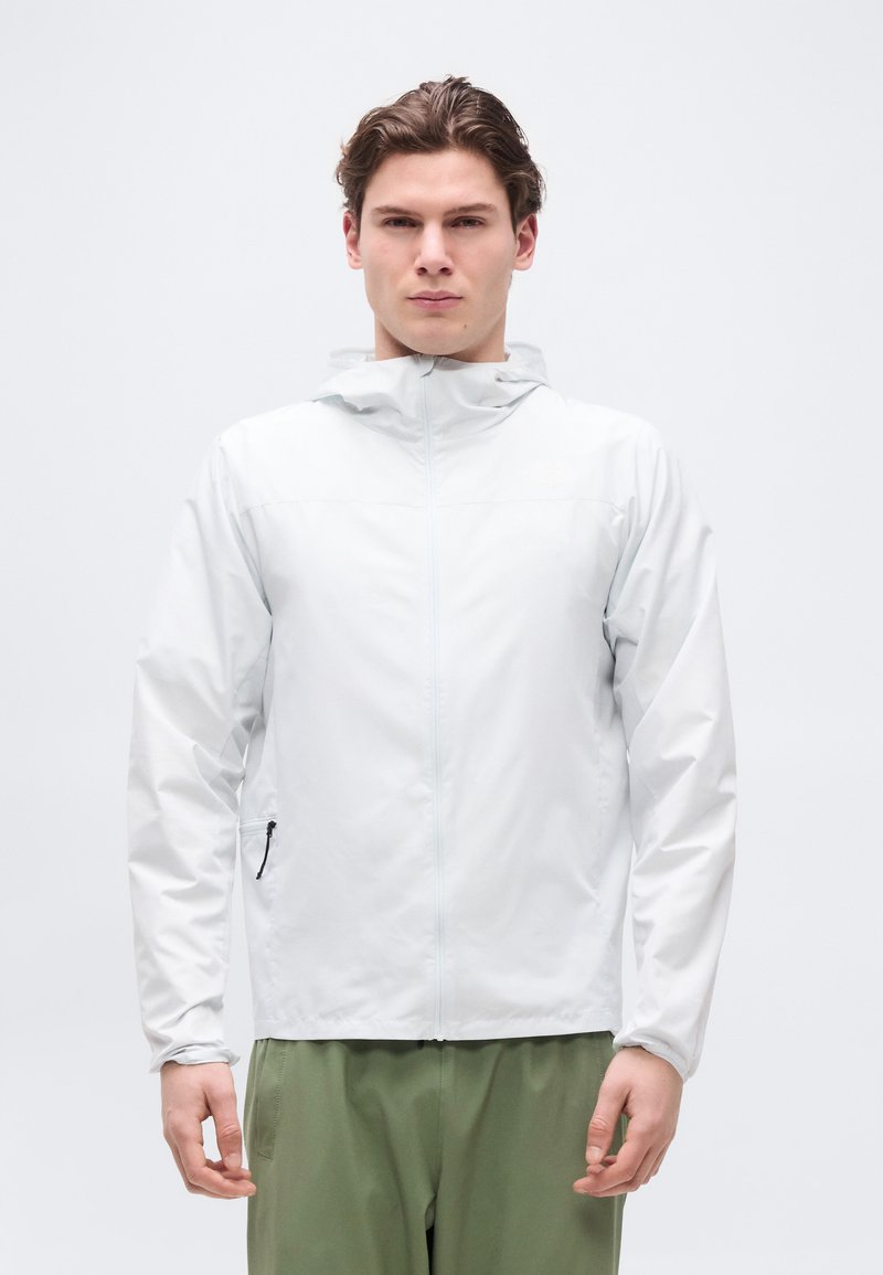 Νεαρός άνδρας που φοράει ανοιχτό γκρι μπουφάν με κουκούλα και φερμουάρ και ελιά πράσινο παντελόνι, στέκεται μπροστά σε λευκό φόντο.