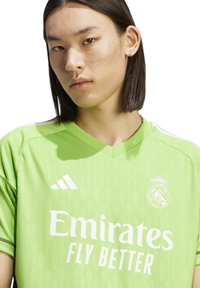 Camiseta de fútbol verde con "Emirates" y "Fly Better" en blanco. Diseño de cuello en V con patrones de diamantes en relieve y logo de adidas.