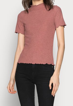 Vrouw draagt een aansluitende stoffige roze geribbelde top met korte mouwen en een krulzoom, en zwarte hoge taille broek tegen een effen achtergrond.