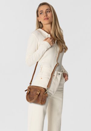 Sac en bandoulière en cuir marron avec une texture lisse, de forme rectangulaire et avec des finitions dorées. Porté avec un haut léger à manches longues et un jean blanc.