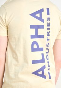 Krém színű póló, amelyen egy nagy, függőleges grafika található sötétkék színben, amelyen az "ALPHA INDUSTRIES" felirat szerepel. Puha, sima pamut anyag.
