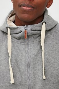 Siva zip-hoodie s mekim teksturom, s visokim ovratnikom, kremasto-bijelim špagicama i suptilnom oznakom na patentnom zatvaraču.