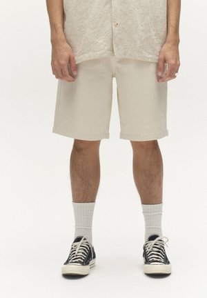 Bărbat care poartă pantaloni scurți bej până la genunchi, șosete albe cu dungi, adidași negri și o cămașă bej cu textură, stând în fața unui fundal alb.