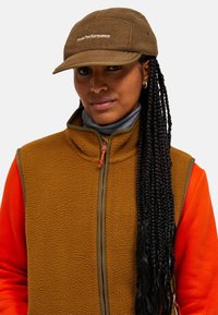 Braune Fleece-Weste mit orangefarbenen Ärmeln, hohem Kragen und Frontreißverschluss. Beiger Hut mit gesticktem "Peak Performance"-Logo. Lange geflochtene Haare.