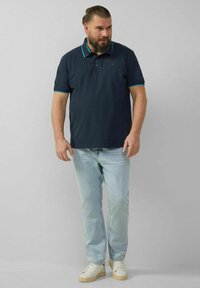 Marineblaues Poloshirt mit türkisfarbenen Akzenten, kurzen Ärmeln und Kragen, kombiniert mit hellblauen Jeans und weißen Sneakers.