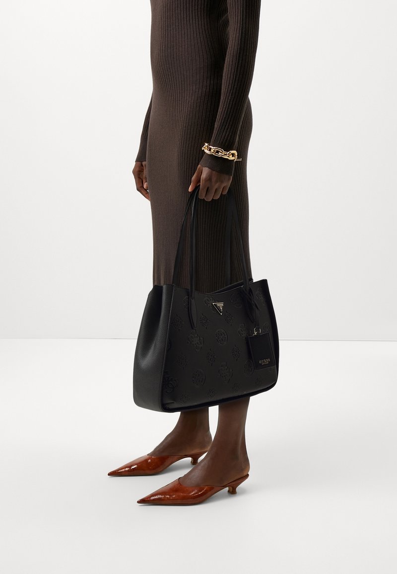 Personne en robe marron côtelée tenant un sac cabas noir à motifs, portant des chaussures marron pointues à petit talon et un bracelet chaîne doré.