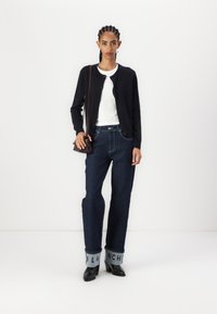 Cardigan negro, top blanco de canalé, jeans de pierna ancha oscuros con puños azul claro que presentan el texto "BLANCH", y botas de tacón negras.