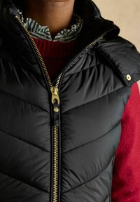 Czarna kamizelka puchowa w szewronowy wzór, z złotym zamkiem błyskawicznym i napami. Nosi się ją na czerwonym swetrze i zielono-paskowanej koszuli z kołnierzykiem.