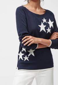 Femme portant un pull bleu marine avec des motifs d'étoiles blanches sur le devant et les manches, associée à un pantalon blanc, bras croisés.