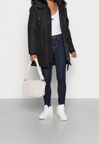 Musta parkatakki tekoturkisvuorella, valkoinen olkalaukku punotulla tekstuurilla, tummansiniset kapeat farkut ja valkoiset korkeavartiset tennarit.