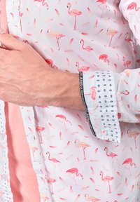 Witte shirt met een patroon van roze flamingo's en veren; heeft een contrasterende azuurblauwe stippenmanchet en een roze verticale streep aan één kant.
