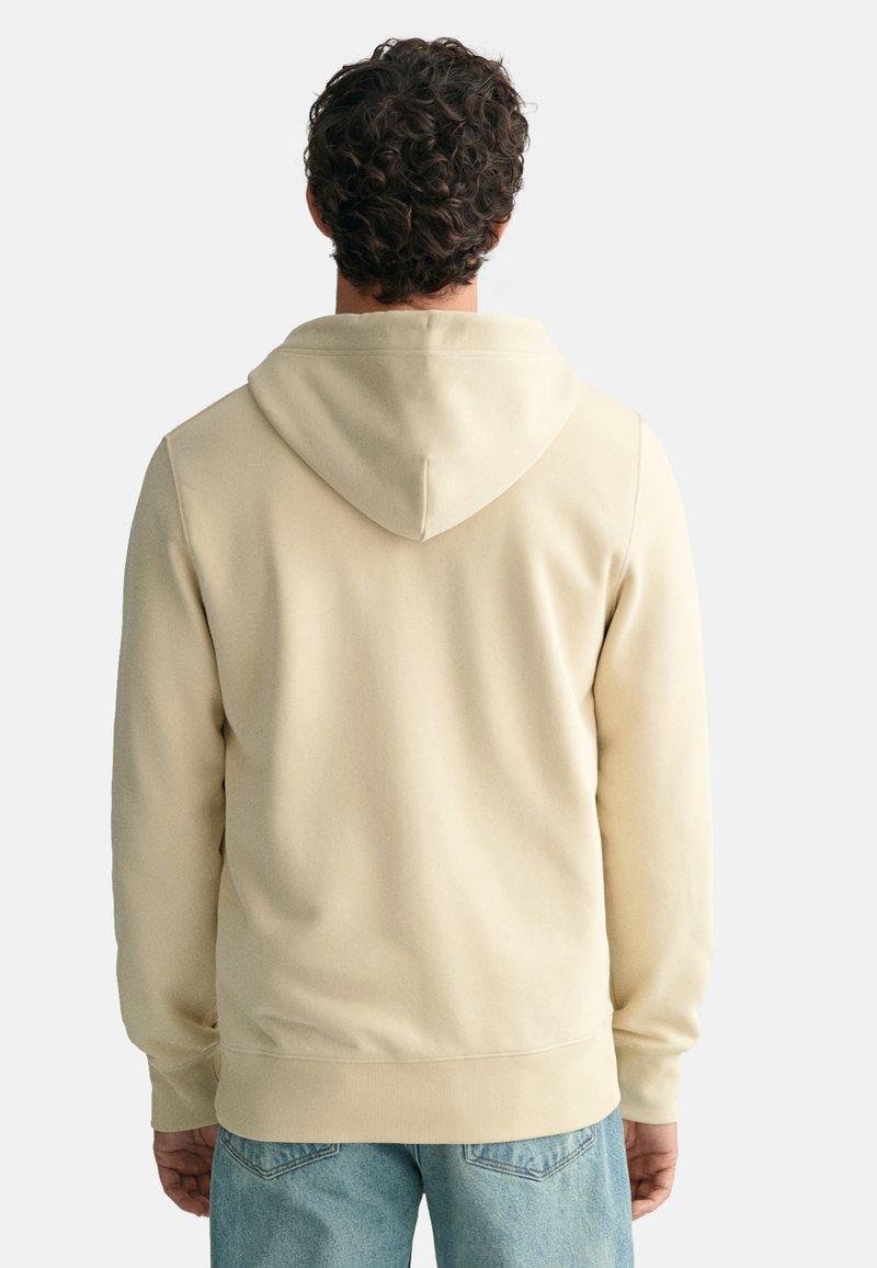 Beige Kapuzenpullover mit einer weichen Textur, elastischen Bündchen und einem lockeren Schnitt, von hinten vor einem schlichten Hintergrund betrachtet.