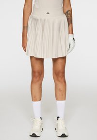 Plisserad beige golfkjol med hög midja som har en svart logotyp. Buren med vita strumpor och ljusa sportskor, stående mot en enkel bakgrund.